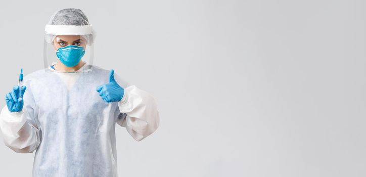 Covid-19, medical research, healthcare workers and quarantine concept. Confident professional doctor in personal protective equipment PPE costume, show thumb-up, hold syringe with corona vaccine.