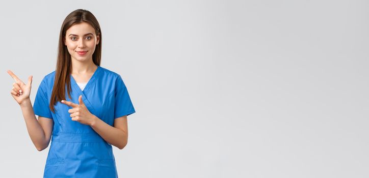 Healthcare workers, prevent virus, insurance and medicine concept. Smiling attractive female doctor or nurse in blue scrubs pointing fingers left to show patients advertisement, important info.