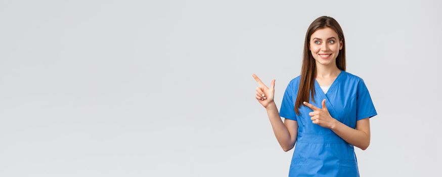 Healthcare workers, prevent virus, insurance and medicine concept. Smiling pretty nurse or doctor in blue scrubs, pointing fingers left and looking at baner with pleased grin, grey background.