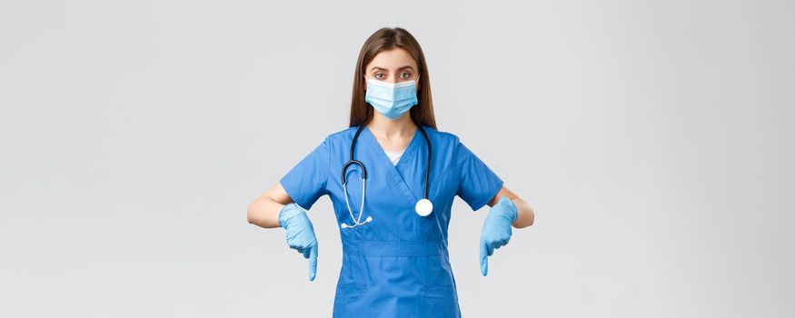 Covid-19, preventing virus, health, healthcare workers and quarantine concept. Determined female nurse or doctor in blue scrubs, medical mask and gloves, pointing fingers down, showing info banner.