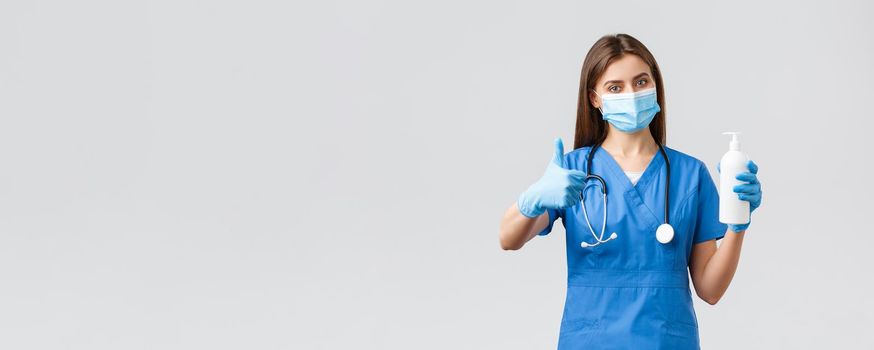 Covid-19, preventing virus, health, healthcare workers and quarantine concept. Confident female doctor, nurse in blue scrubs in medical mask and scrubs advice using hand sanitizer, thumbs-up approval.