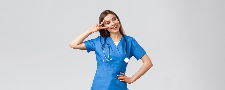 Medical workers, healthcare, covid-19 and vaccination concept. Cheerful attractive female nurse or doctor in blue scrubs, stethoscope, show peace sign, stay optimistic, standing grey background.