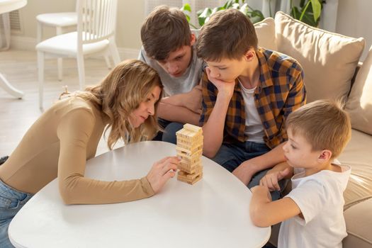 Three boys and woman, in a sunny room, are enthusiastically playing a board game made of wooden rectangular blocks, pulling pieces out of the tower. Top view
