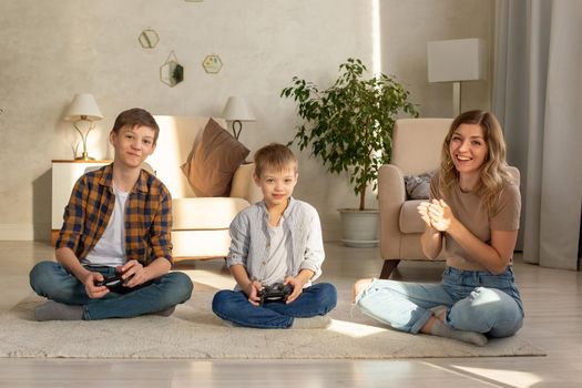 Portrait of two smiling boys and happy woman, sits on the floor in a room playing video games with joysticks. Close up
