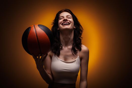 Teenage girl with dental braces holding basketball. Studio portrait on orange colored background.