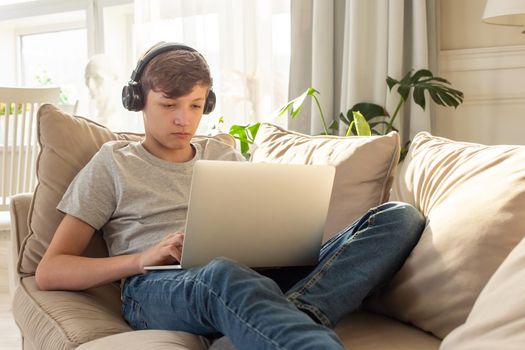 Portrait of teenager boy in a gray t-shirt and blue jeans, lying on a sofa in a room, wearing black headphones on his head, looks into a gray laptop, on his knees. Copy space