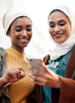 Cropped shot of two attractive young women wearing headscarves and standing together while using a cellphone in the city.