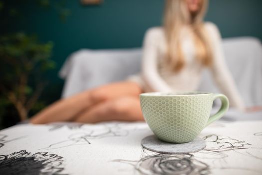 A young sexy woman with shapely legs sits on a sofa and a cup of aromatic coffee stands in front of her.