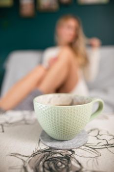 A young sexy woman with slim legs sits on a living room sofa and a porcelain cup with aromatic coffee stands in front of her.