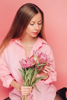 A sweet charming woman with flowers in a pink dress on a pink background smiles, happiness and luck.