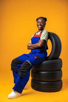 Portrait of smiling black female mechanic posing with new car tyres in yellow studio