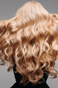 Back view of unrecognizable woman in black with volumous, blonde wavy hair over grey background. Isolated on grey. Studio. Perfect healthy hair. Hairdresser.