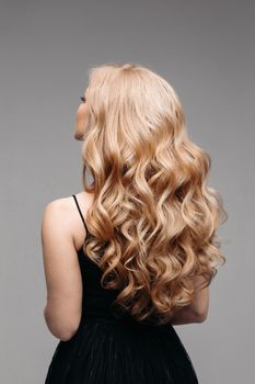 Back view of unrecognizable woman in black with volumous, blonde wavy hair over grey background. Isolated on grey. Studio. Perfect healthy hair. Hairdresser.