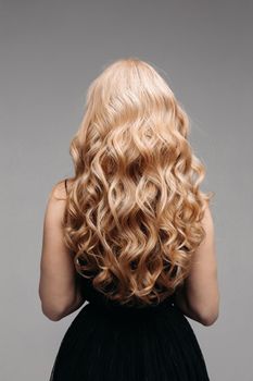 Back view of unrecognizable woman in black with volumous, blonde wavy hair over grey background. Isolated on grey. Studio. Perfect healthy hair. Hairdresser.