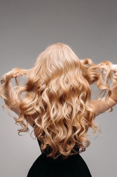 Back view of unrecognizable woman in black with volumous, blonde wavy hair over grey background. Isolated on grey. Studio. Perfect healthy hair. Hairdresser.