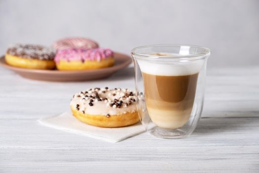 Glazed decorated donuts on plates and coffee latte or cappuccino with milk foam in a heat-resistant glass cup. Selective focus.