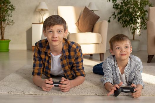 Portrait of two smiling boys lie on the floor in a room playing video games with joysticks. Close up