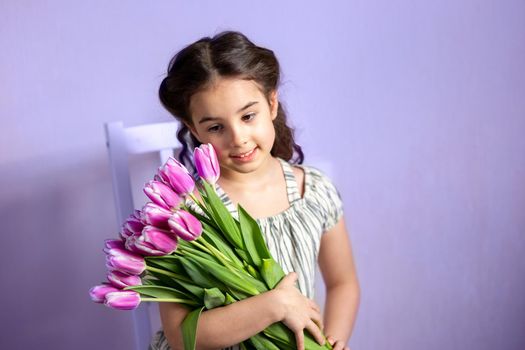 Beautiful little girl with a bouquet of tulips, sits on a white chair, against a purple wall, smiles. Copy space