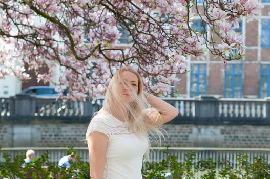Beautiful young woman with long hair looking up at blooming tree with pink magnolia flowers.