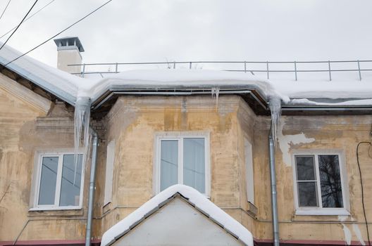 Multiple ice icicles hang from a drainpipe at the edge of the roof. Against the background of the wall of an old brick house. Large cascades, even beautiful rows. Cloudy winter day, soft light.