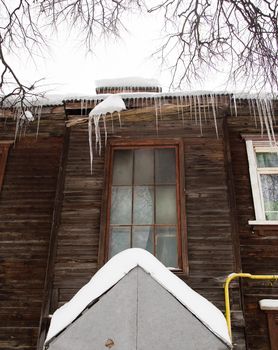 Winter transparent icicles hang on the edge of the roof. Against the background of the wooden wall of the old house. Large cascades, even beautiful rows. Cloudy winter day, soft light.