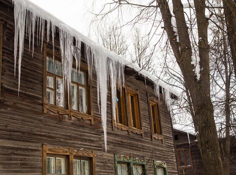 Frozen transparent icicles hang on the edge of the roof. Against the background of the wooden wall of the old house. Large cascades, even beautiful rows. Cloudy winter day, soft light.