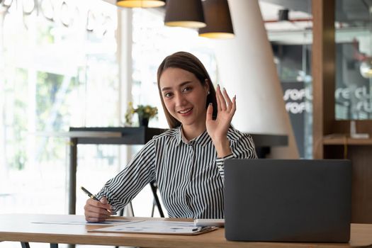 happy asian woman smiling and waving hand at laptop, while speaking or chatting on video call in office. online conference webinar concept