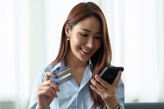 Young beautiful Asian woman using smartphone and credit card for online shopping at home with copy space. E-payment technology, shopaholic lifestyle, or mobile phone financial application concept.