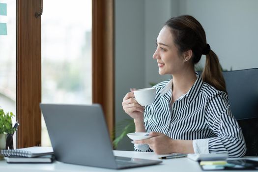 Beautiful young Asian businesswoman smiling holding a coffee mug and laptop working at the office.