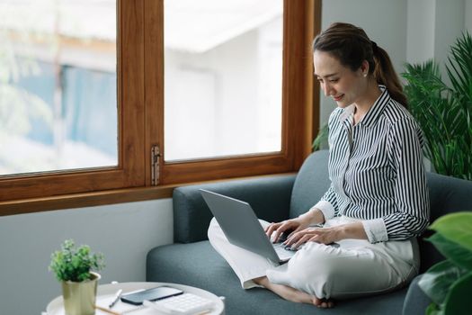 Asian young woman working from home office sit on couch hold paper invoice check information online.