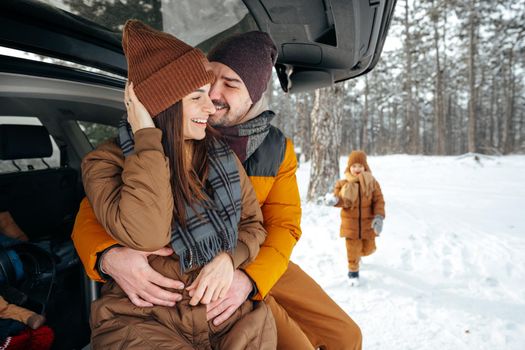 Lovely smiling couple sitting in car trunk in winter forest, close up