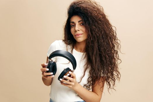 Fashionable Latin woman with afro ready to enjoying cool songs, hold wireless headphones, close eyes, stay isolated near beige wall. Copy free space for design ad. Fashion IT Seasonal Sale concept