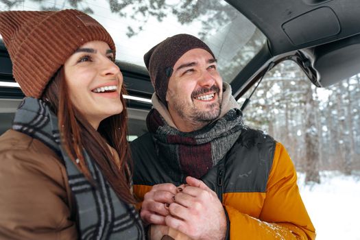 Lovely smiling couple sitting in car trunk in winter forest, close up