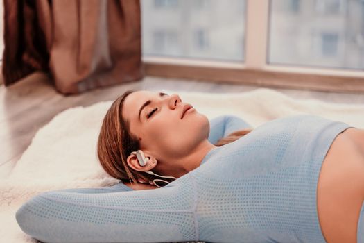 Side view portrait of relaxed woman listening to music with headphones lying on carpet at home. She is dressed in a blue tracksuit