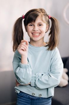 Portrait of cute little child girl is rejoices and showing her lost milk tooth and smiling to camera of toothless mouth while she standing in bedroom. Happy girl holding her fallen tooth in hand