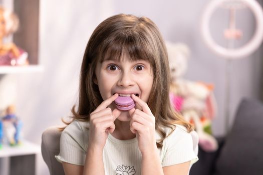 Young fresh and beautiful brunette kid girl smiles, thinks about something, looks on small macaron cookie in her hand and bites it.