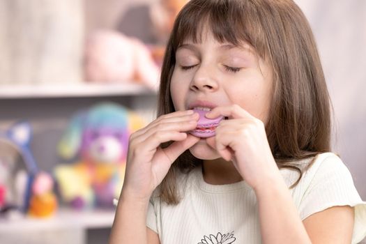 Attractive brunette school girl on a home background, licks her lips, eating one violet macaron, closes her eyes with pleasure. Dessert person, sweet tooth, gourmet. Feeling good, delicious.