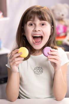 Hilarious school girl wants to eat two macarons at the same time, widely opens her mouth. Dessert, delicious. Tasty sweet color macaron. Feeling good, delicious.