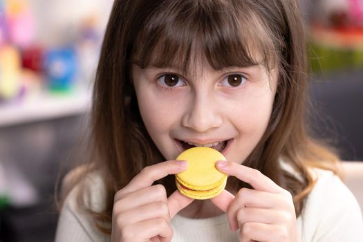 Brunette school girl on a home background, licks her lips, eating one yellow macaron. Dessert person, sweet tooth, gourmet. Feeling good, delicious.