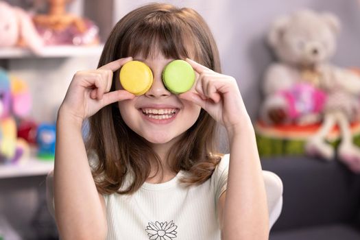 Happy smiling face of a little girl covering her eyes with macaroons on a colored home background. Sweet tooth, unhealthy food, no diet. Being happy. Female portrait