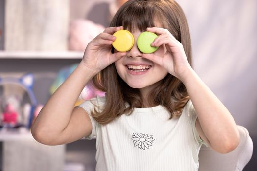 Teen girl plays with dessert macarons, holding the cookies like glasses around the eyes and throws his hands to the sides. Happy smiling face of little girl covering her eyes with macaroons.