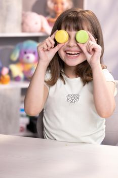 Happy smiling face of little girl covering her eyes with macaroons. Teen girl plays with dessert macarons, holding the cookies like glasses around the eyes and throws his hands to the sides.
