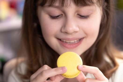 Attractive brunette school girl on a home background, licks her lips, eating one yellow macaron, closes her eyes with pleasure. Dessert person, sweet tooth, gourmet. Feeling good, delicious.