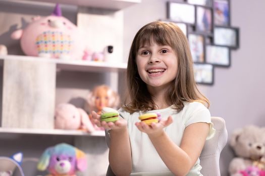 Attractive smiling cute school kid girl holds four tasty cookies in her hands and look at camera. Traditional French multicolored macaroon. Food concept.