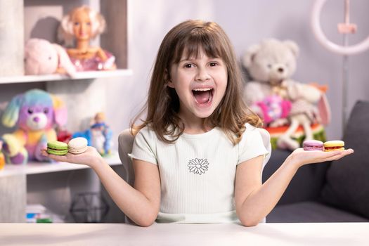 Attractive smiling cute school kid girl holds four tasty cookies in her hands and look at camera. Traditional French multicolored macaroon. Food concept.