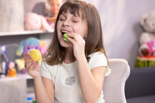 Young brunette kid girl can not make right decision and choice what macaron to bite first - left from with lemon and beryy filling or right made with pistachio cream, then bite macaron in green color.