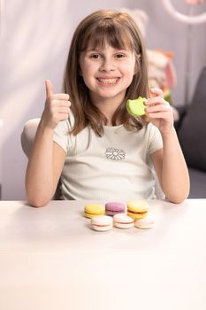 Attractive young scholl girl bites a macaroon, surprised with taste, cheerfully smiles shows a finger up. Positive emotions, being a gourmet, sweet tooth, delicious. Female portrait on home background