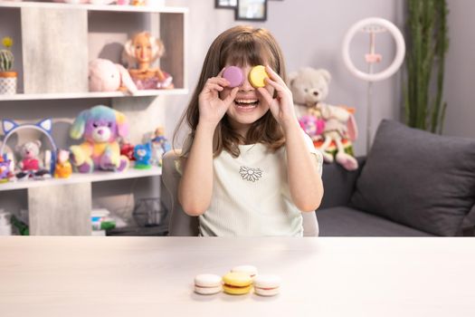 Teen girl plays with dessert macarons, holding the cookies like glasses around the eyes and throws his hands to the sides. Happy smiling face of little girl covering her eyes with macaroons.