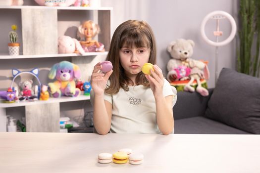 Young brunette kid girl can not make right decision and choice what macaron to bite first - right from with lemon and beryy filling or left made with violet cream, then bite macaron in yellow color.