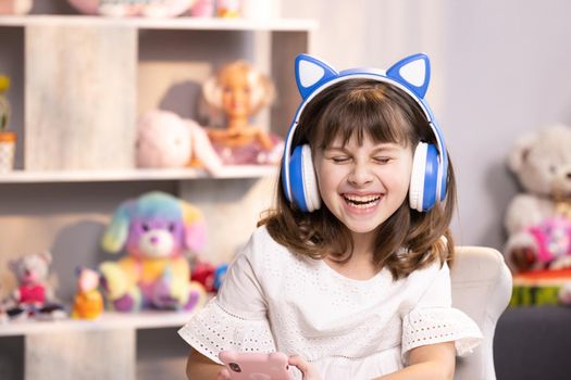 Cute small kid girl in headphones using funny editing application on smartphone, enjoying cool video or music content in social network, playing online games, communicating distantly.